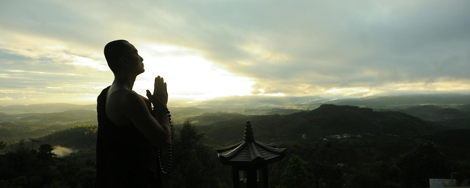 A monk in silhouette prays peacefully at sunrise with mountains in the background.